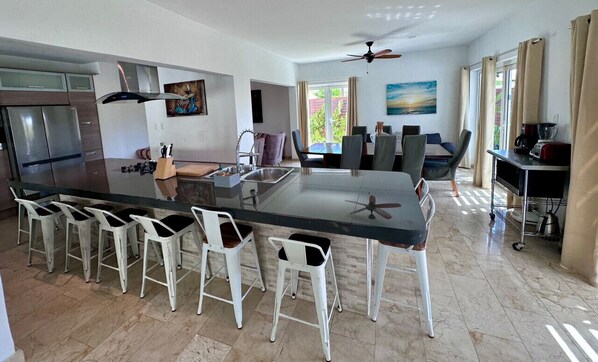 Een moderne keuken met een centraal eiland, barkrukken, een eethoek met stoelen, een koelkast en een plafondventilator.