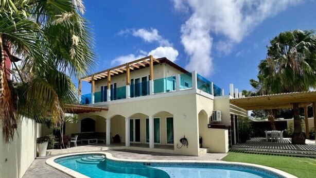 A two-story house with a swimming pool, surrounded by palm trees and a clear blue sky.