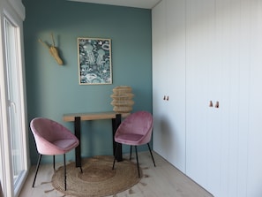 Ein Zimmer mit türkisfarbenen Wänden, einem Holztisch, zwei rosafarbenen Stühlen, einem gewebten Teppich, einer Wanddekoration und einem weißen Kleiderschrank.