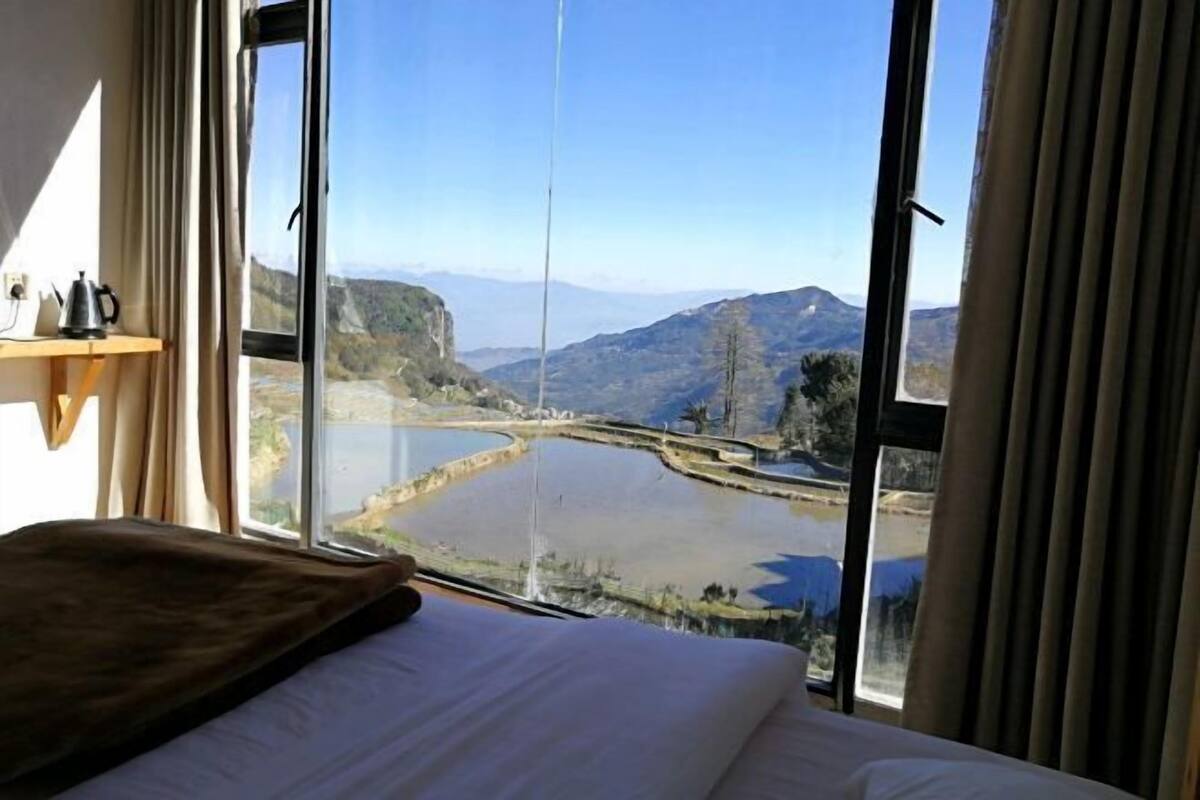 A bedroom with a large window offering a view of a river and mountains.