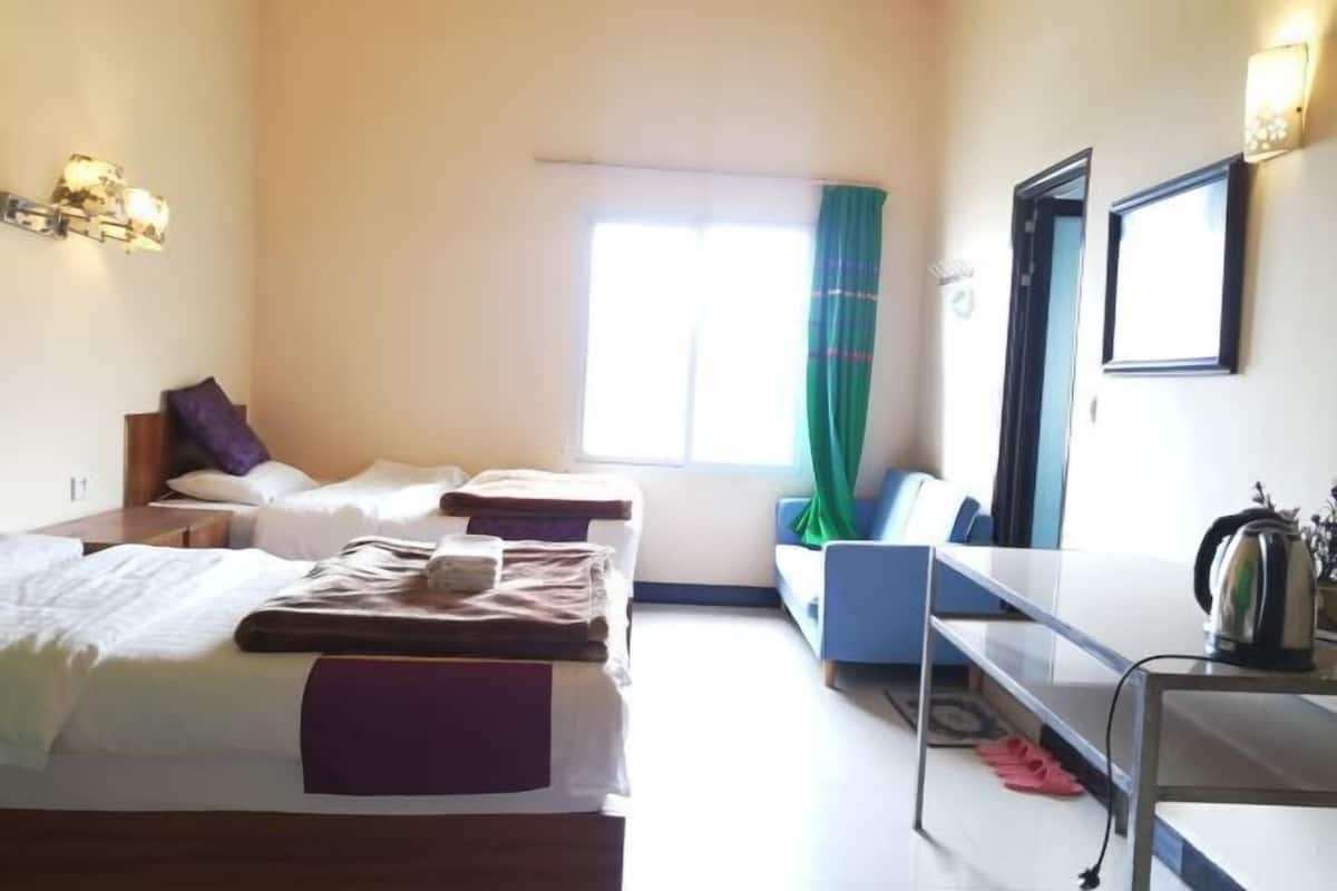 A hotel room with two beds, a desk, a chair, a window with curtains, and a wall-mounted light fixture.