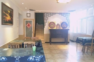 Un comedor con una mesa larga, sillas y un mantel estampado. Se ve una escalera que sube a un nivel superior y una puerta con un felpudo decorativo.