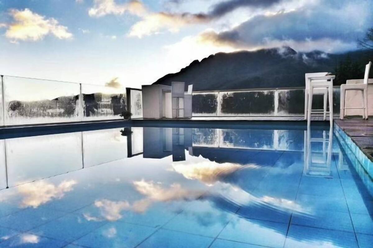 A swimming pool with a clear reflection, a covered outdoor area, and a mountain backdrop.