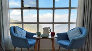 A room with two blue chairs facing a table with a kettle and cups, a large window with a view of hills, and a hanging light fixture. - Jacky's Guesthouse (Yuanyang)