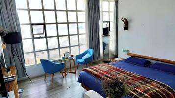 Habitación de hotel con un ventanal grande, una cama con manta escocesa, una silla azul, una mesita con un hervidor y un espejo en la pared.