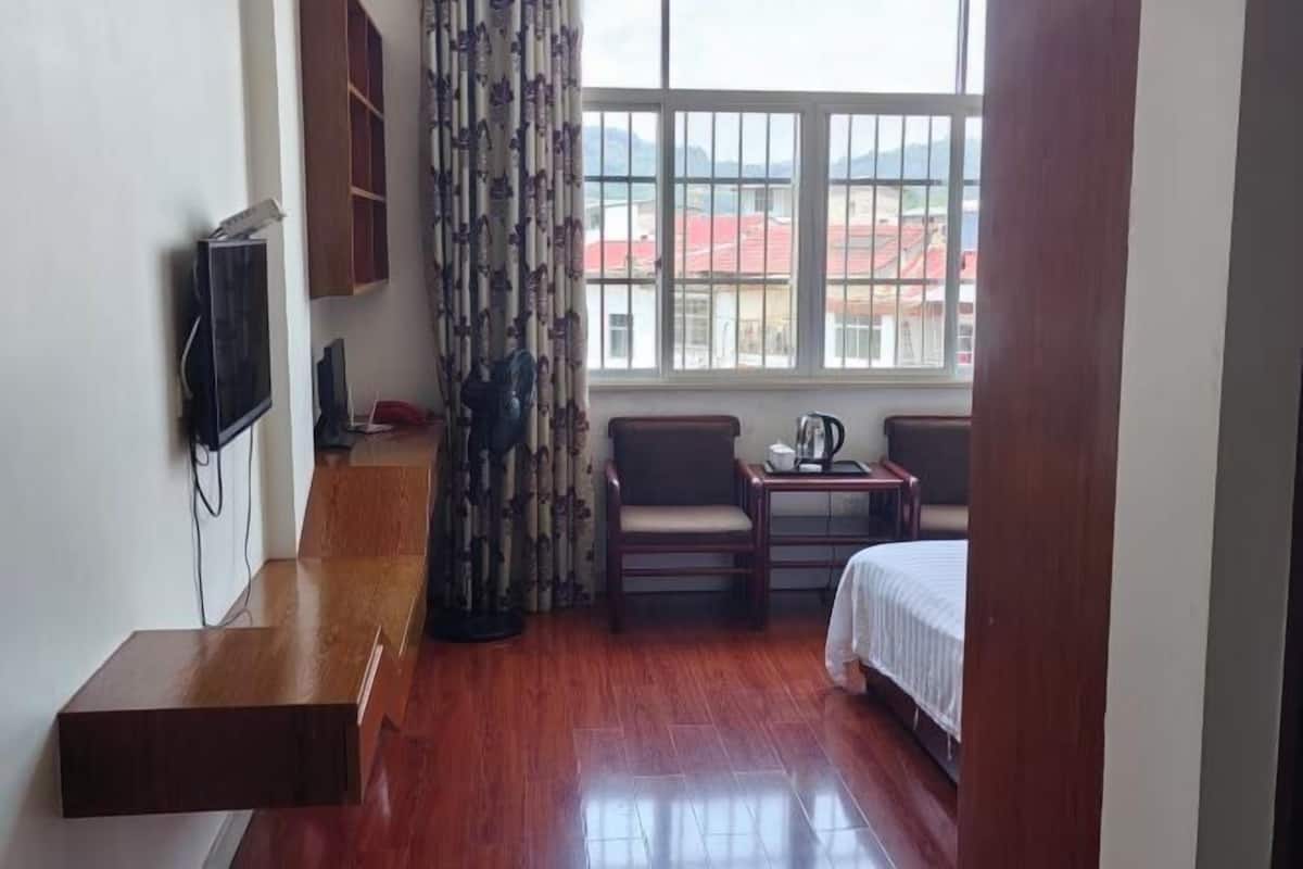 A hotel room with a bed, a wooden bench, a TV, a small table with a kettle, and a window with a view of buildings.
