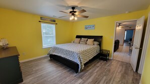 A bedroom with a bed, a ceiling fan, a window with blinds, and a door leading to another room. - Tybee Pink House (Military & Veteran Discount) (Tybee Island)