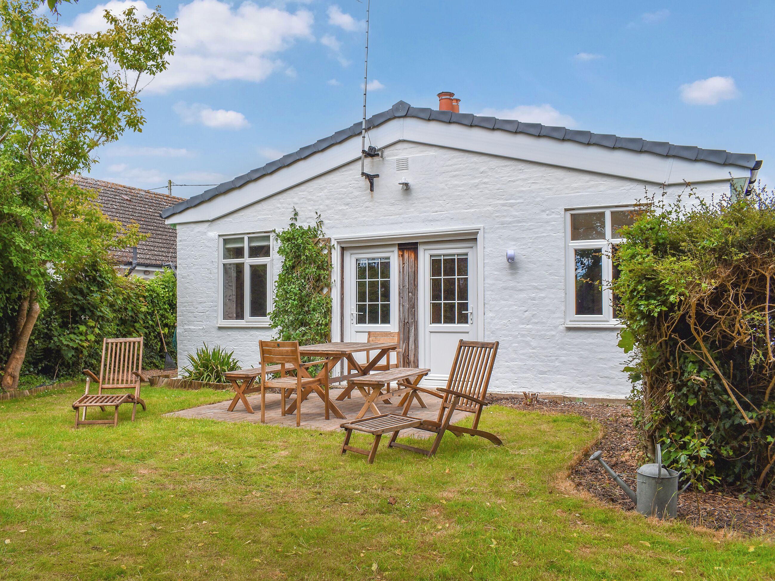 A white house with a tiled roof, a front door, and a small garden with wooden furniture.