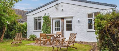 A white house with a tiled roof, a front door, and a small garden with wooden furniture.