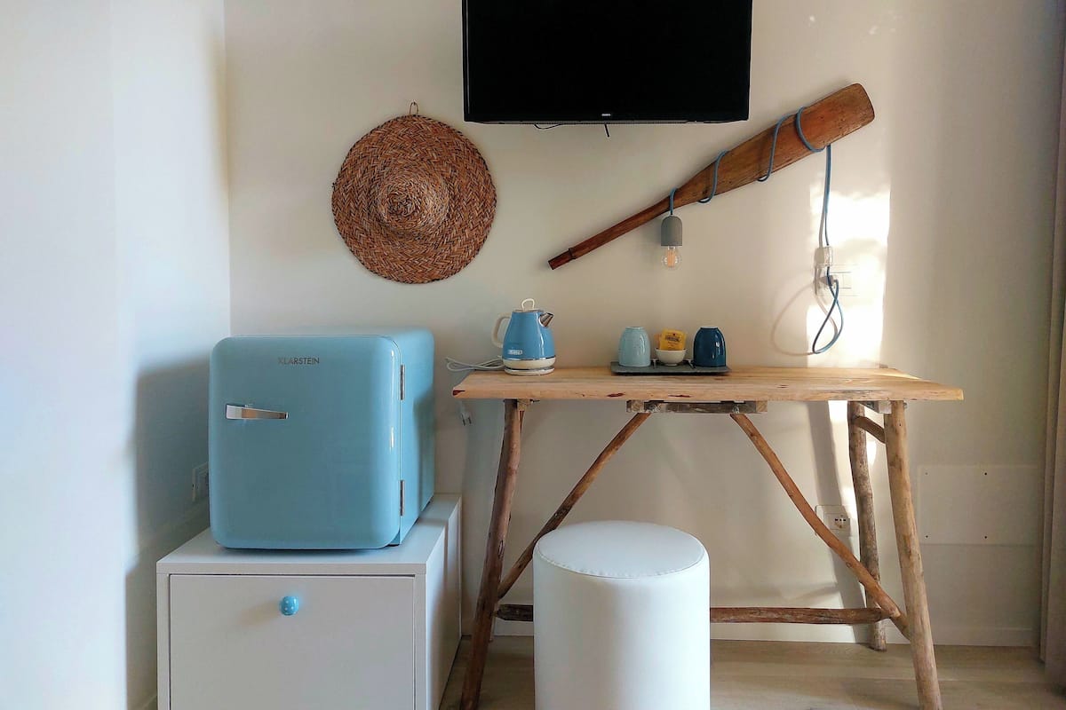 Una habitación con una televisión de pantalla plana en la pared, un refrigerador azul, una mesa de madera con una tetera y tazas, y un reposapiés blanco.