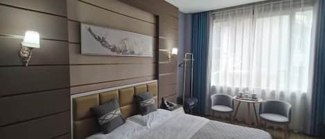 A hotel room with a large bed, a headboard with multiple pillows, a bedside table with a lamp, a small table with chairs, and a large window with curtains.