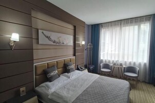 Una habitación de hotel con una cama grande, un cabecero con varios almohadones, una mesita de noche con lámpara, una pequeña mesa con sillas y un ventanal con cortinas.