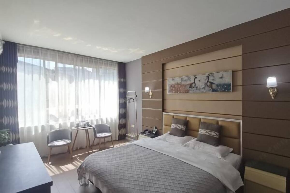 Moderni hotellihuone, jossa on suuri sänky, työpöytä, kaksi tuolia, pieni pöytä ja suuri ikkuna, jossa on verhot.