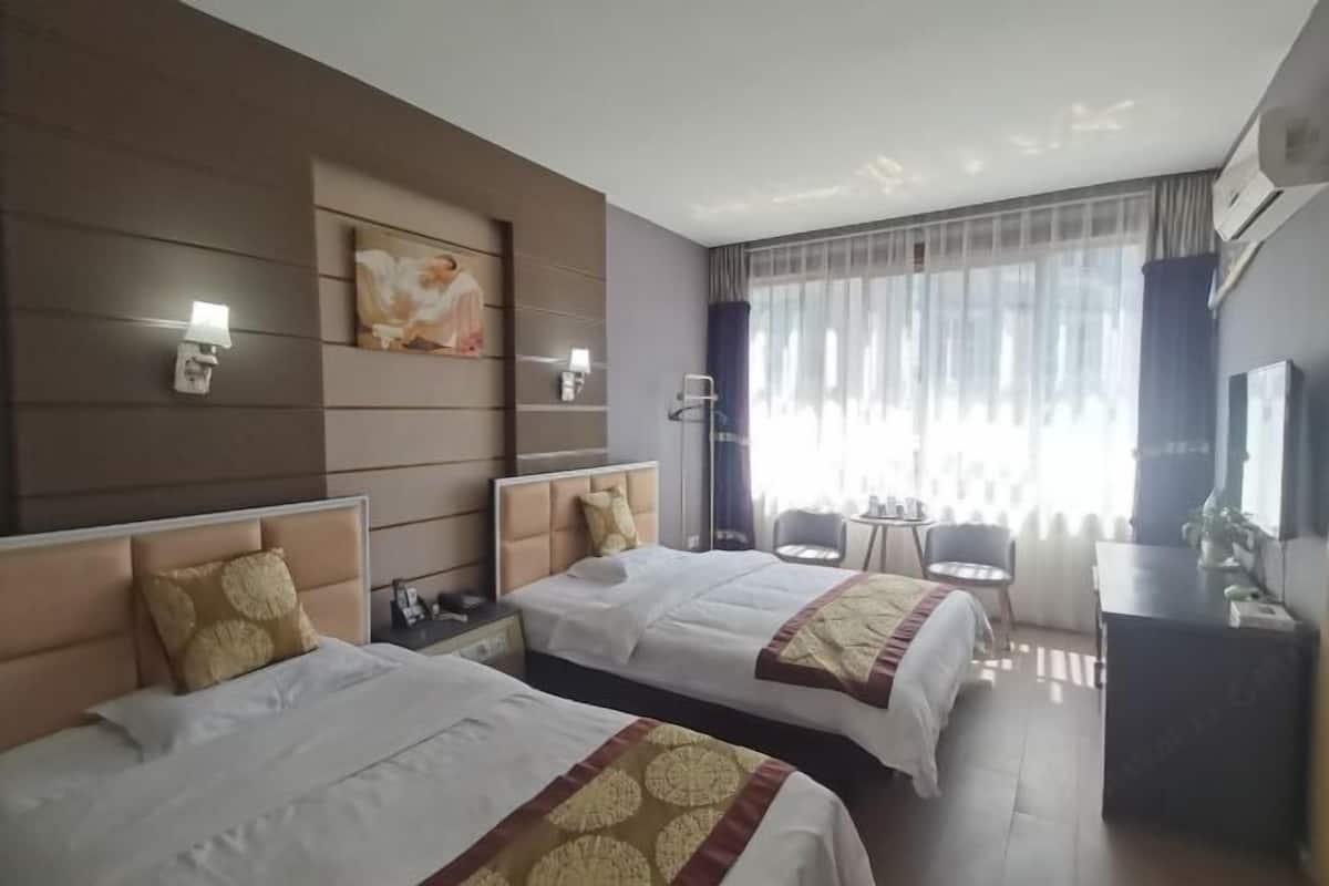 Hotellihuone, jossa on kaksi sänkyä, työpöytä, tuoli, televisio ja suuri ikkuna, jossa on läpikuultavat verhot.