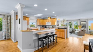 A modern kitchen with wooden cabinets, a central island with bar stools, and a dining area with a sofa and table. - Salish Coast Retreat a High End Home with Views (Port Angeles)