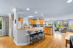 A modern kitchen with wooden cabinets, a central island with bar stools, and a dining area with a sofa and table.