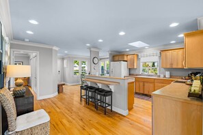 A modern kitchen with wooden cabinets, a central island with bar stools, and a dining area with a sofa and lamp.