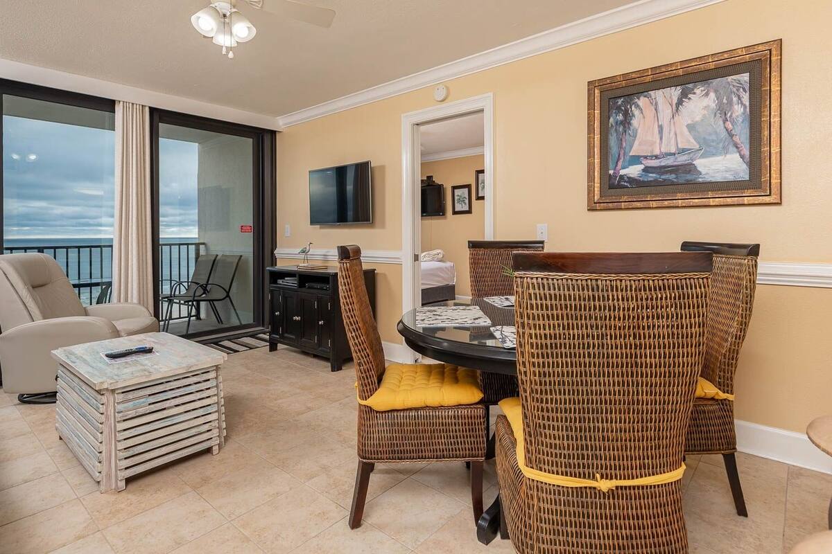 Un balcon spacieux avec vue sur la mer, un coin repas avec des chaises et une table ronde en osier, et un téléviseur fixé au mur.