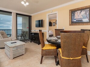 A balcony with a view of the ocean, a dining area with wicker chairs and a table, and a television mounted on the wall. - Island Winds West by Brett/Robinson Vacations (Gulf Shores)