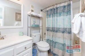 A bathroom with a white sink, a toilet, a shower curtain with a blue and white pattern, and a towel rack with white towels. - Island Winds West by Brett/Robinson Vacations (Gulf Shores)