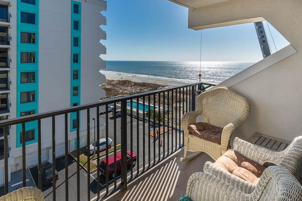 Un balcon doté de mobilier en osier, offrant une vue sur la plage et un espace de stationnement.