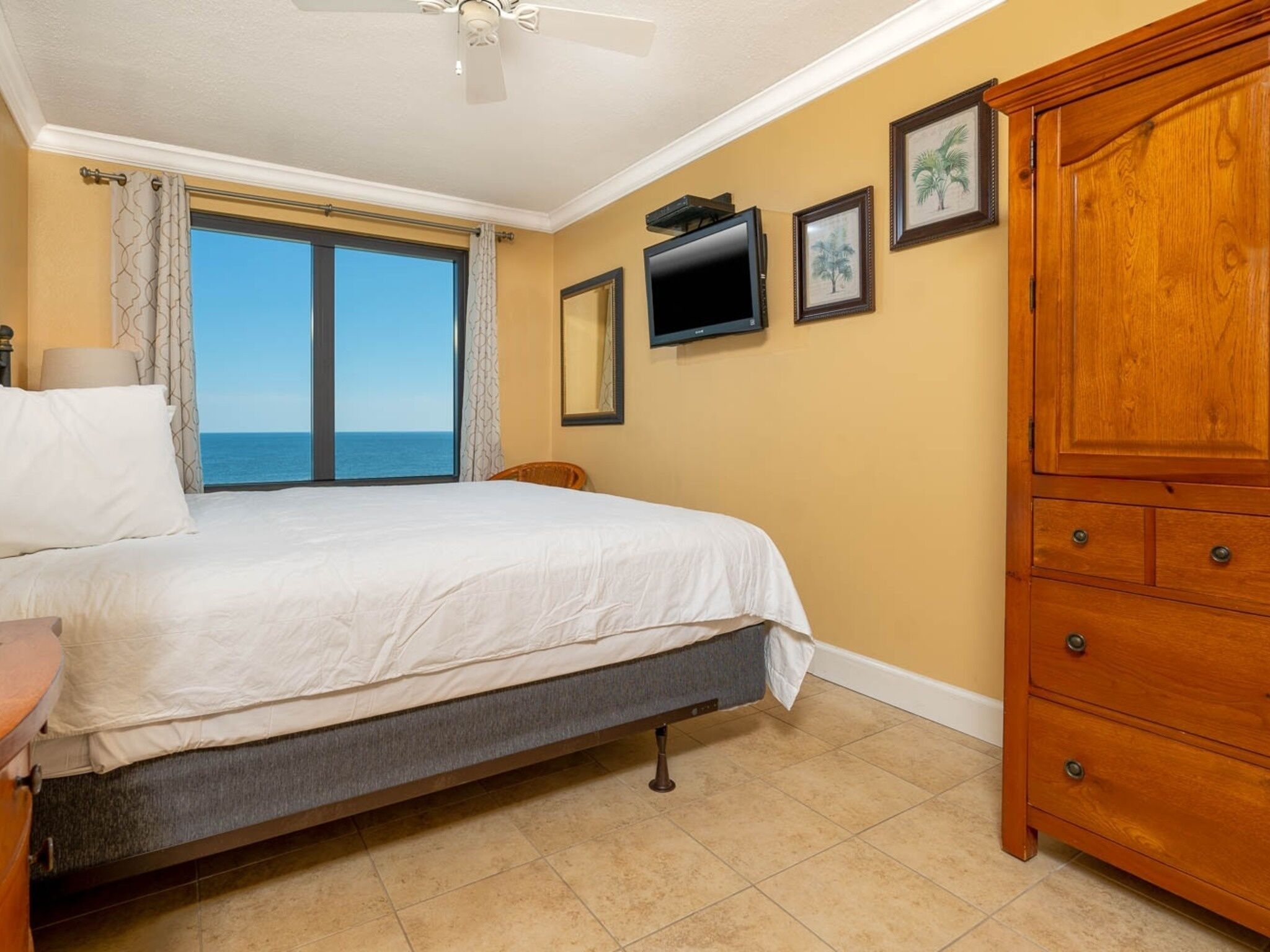 A bedroom with a bed, a wooden dresser, a television, and a view of the ocean.