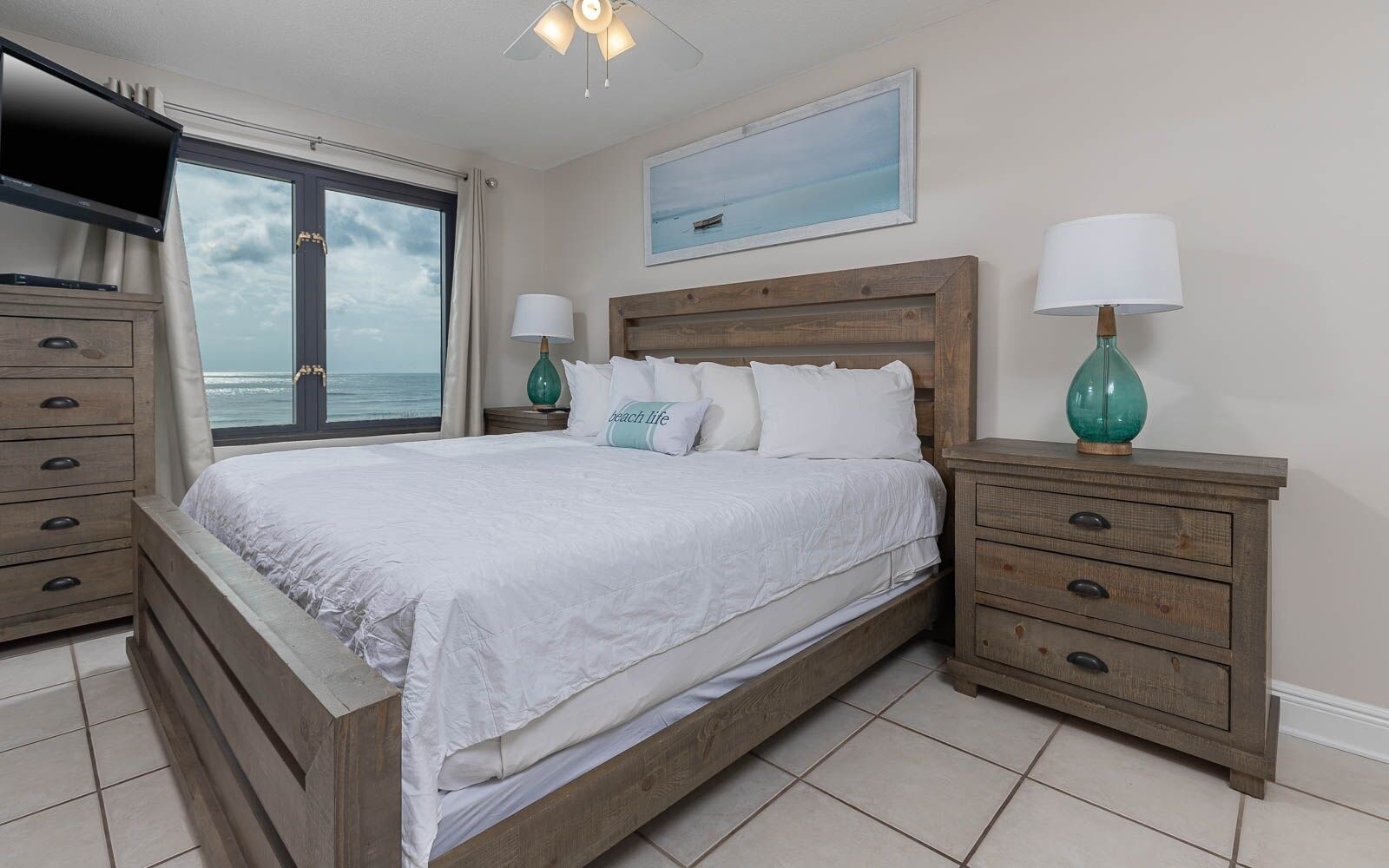 A bedroom with a large bed, wooden headboard, two nightstands with lamps, a dresser, and a view of the ocean through a window.