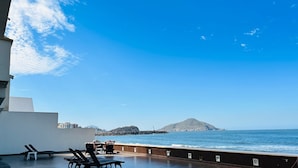 Een terras met loungezetels en een tafel met uitzicht op de zee, met een rotsachtige golfbreker en een berg op de achtergrond.