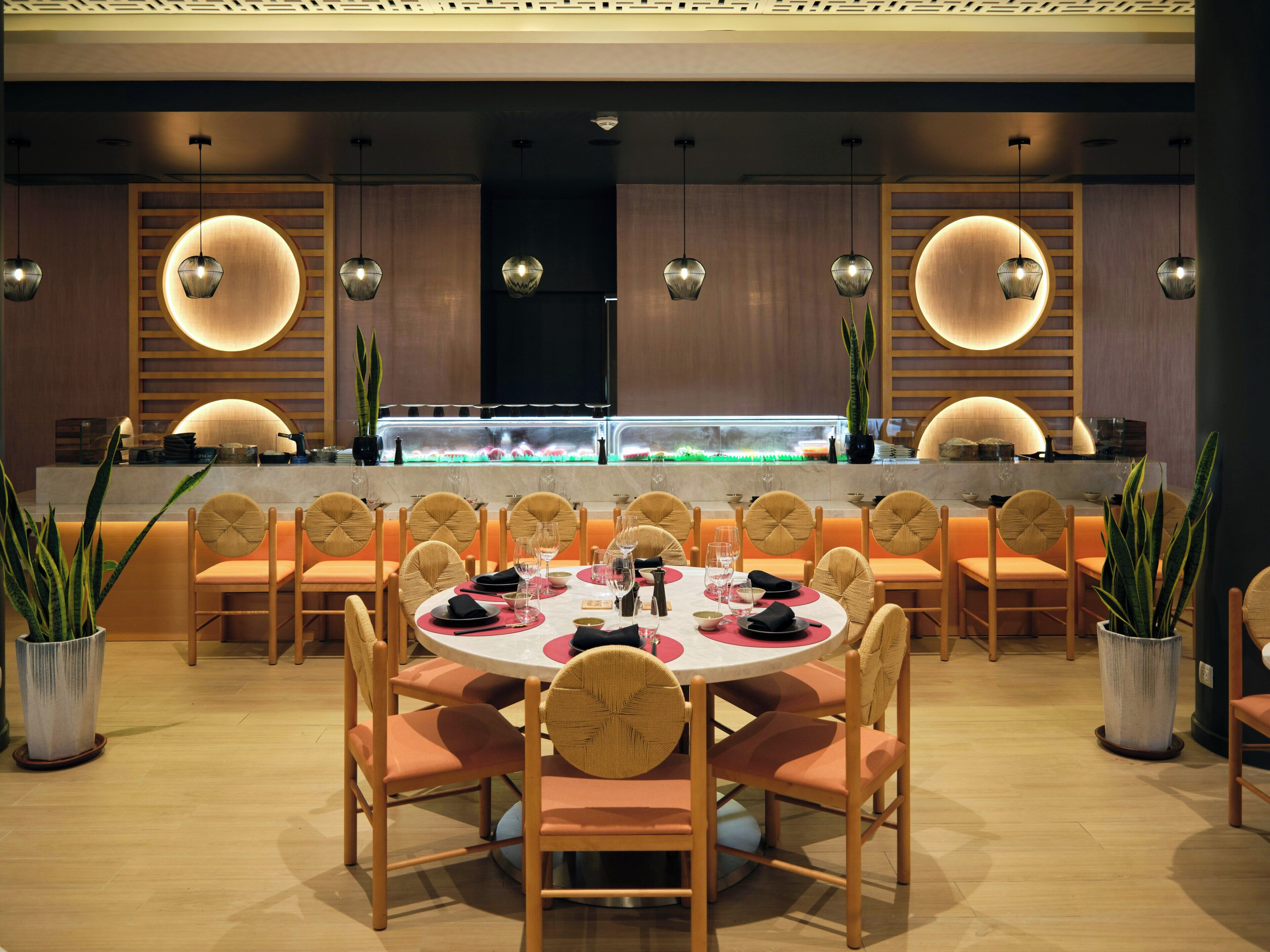 A restaurant with round tables, chairs, pendant lighting, and a sushi bar.