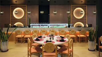 A restaurant with round tables, chairs, pendant lighting, and a sushi bar.