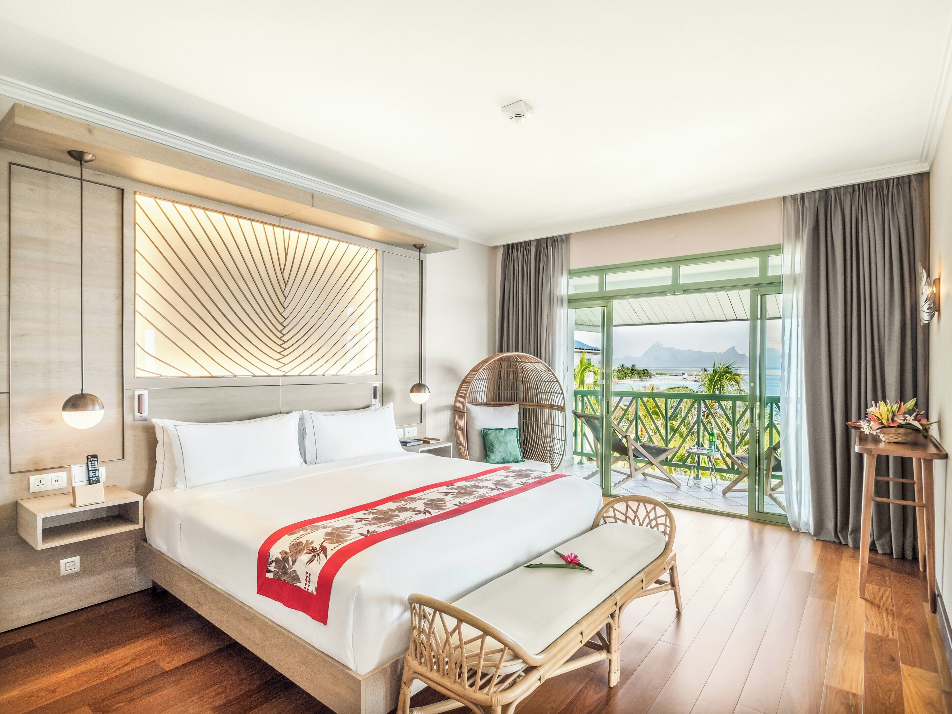 A hotel room with a bed, bedside table, lamp, chair, and a balcony with a view.