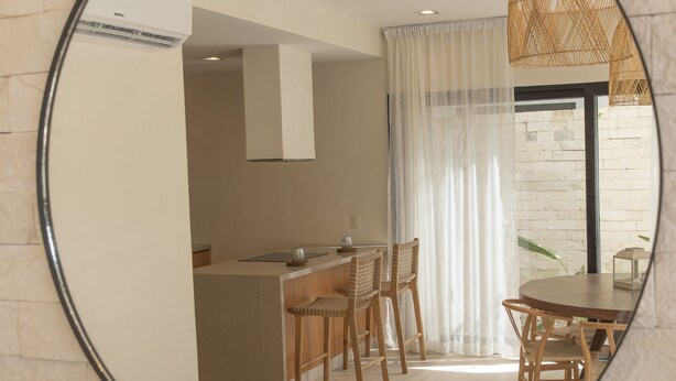 A round mirror reflecting a modern kitchen with a dining area, featuring a stone wall, wooden furniture, and a large window with sheer curtains. - Kuxtal Tulum (Tulum)