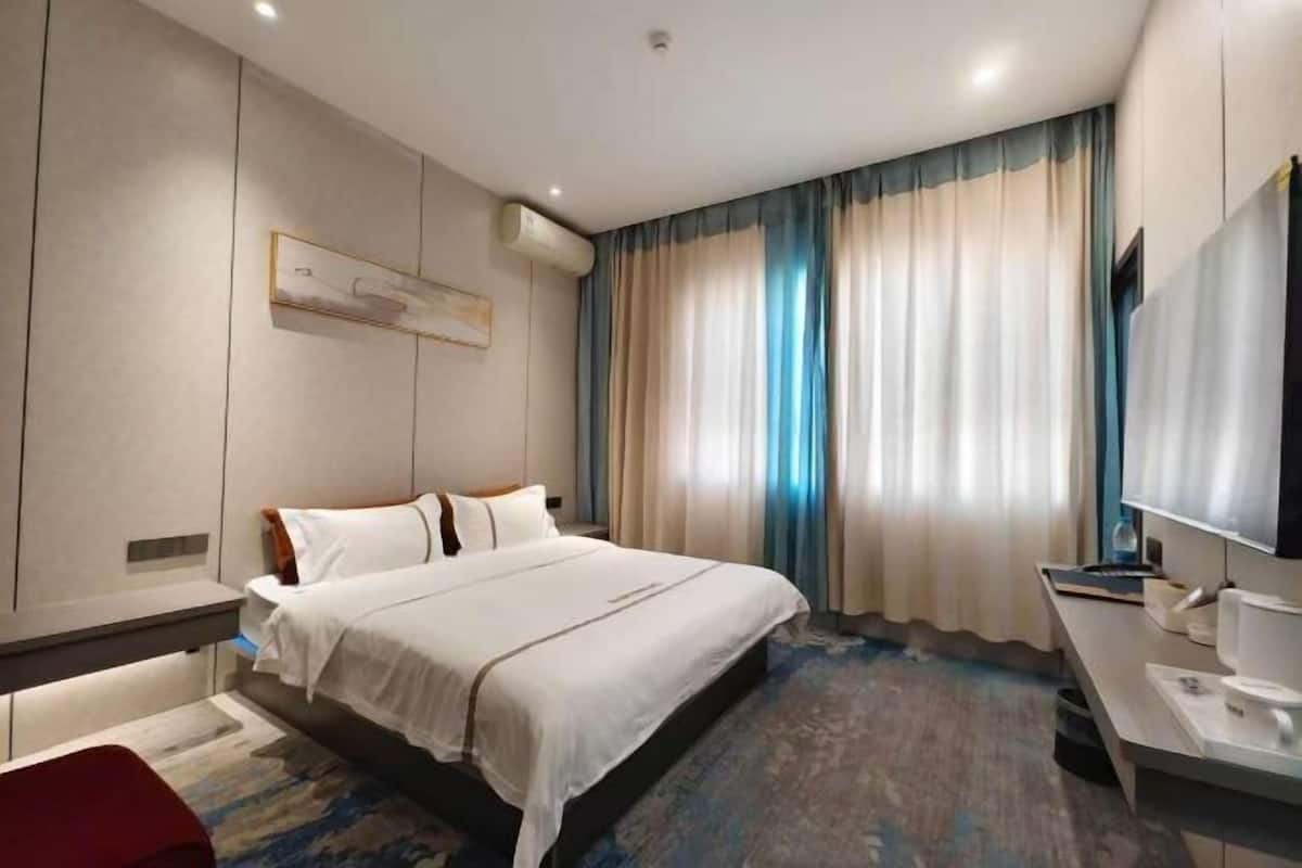 Ein modernes Hotelzimmer mit einem großen Bett, einem Schreibtisch, einem Flachbildfernseher und einem großen Fenster mit Vorhängen.