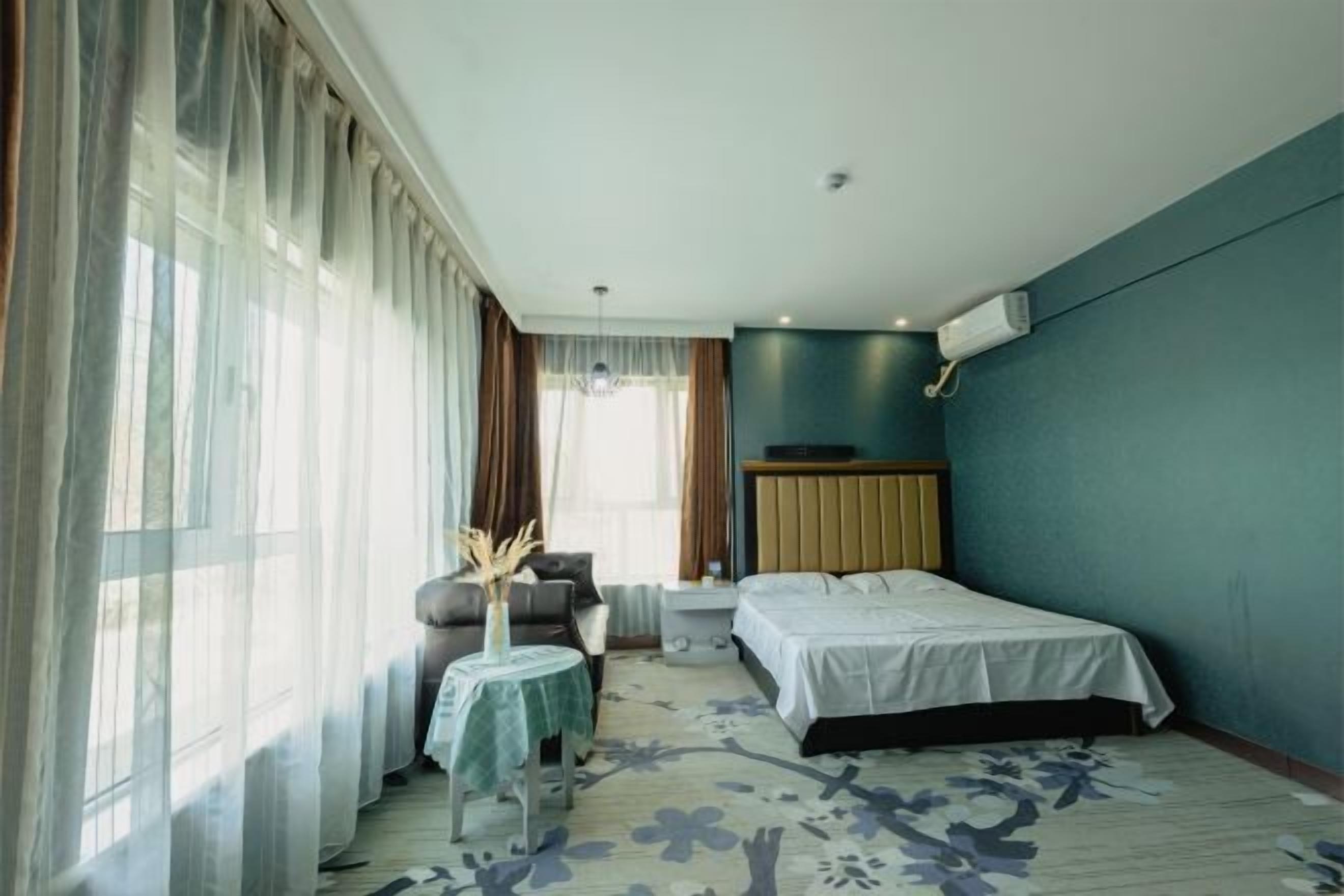 A hotel room with a bed, a small table with a vase, a chair, and a window with curtains.