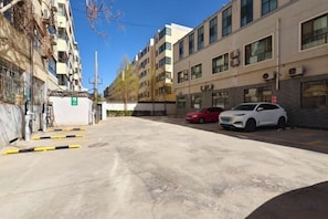Vista da rua com um prédio de vários andares, um carro branco estacionado e um carro vermelho.