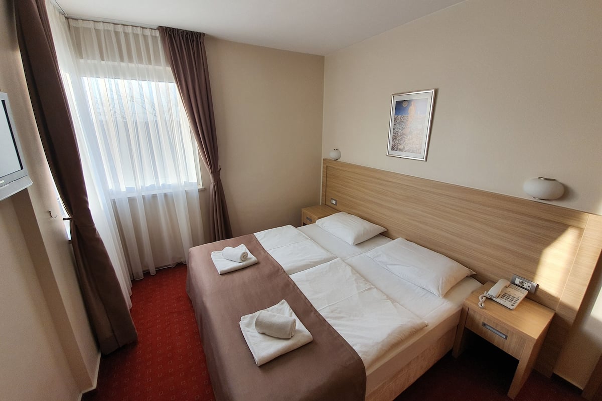 A hotel room with a bed, a bedside table with a lamp, a telephone, and a window with curtains.