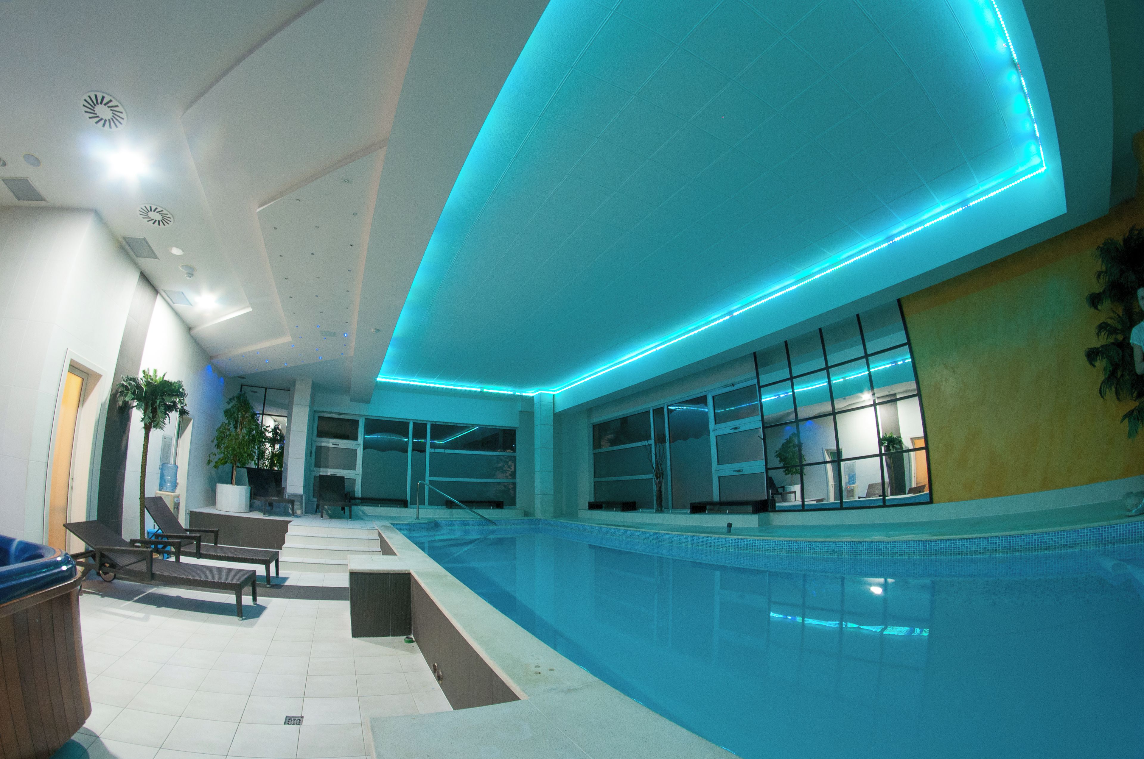 A modern indoor swimming pool with a blue-lit ceiling, clear glass walls, and a seating area with benches.