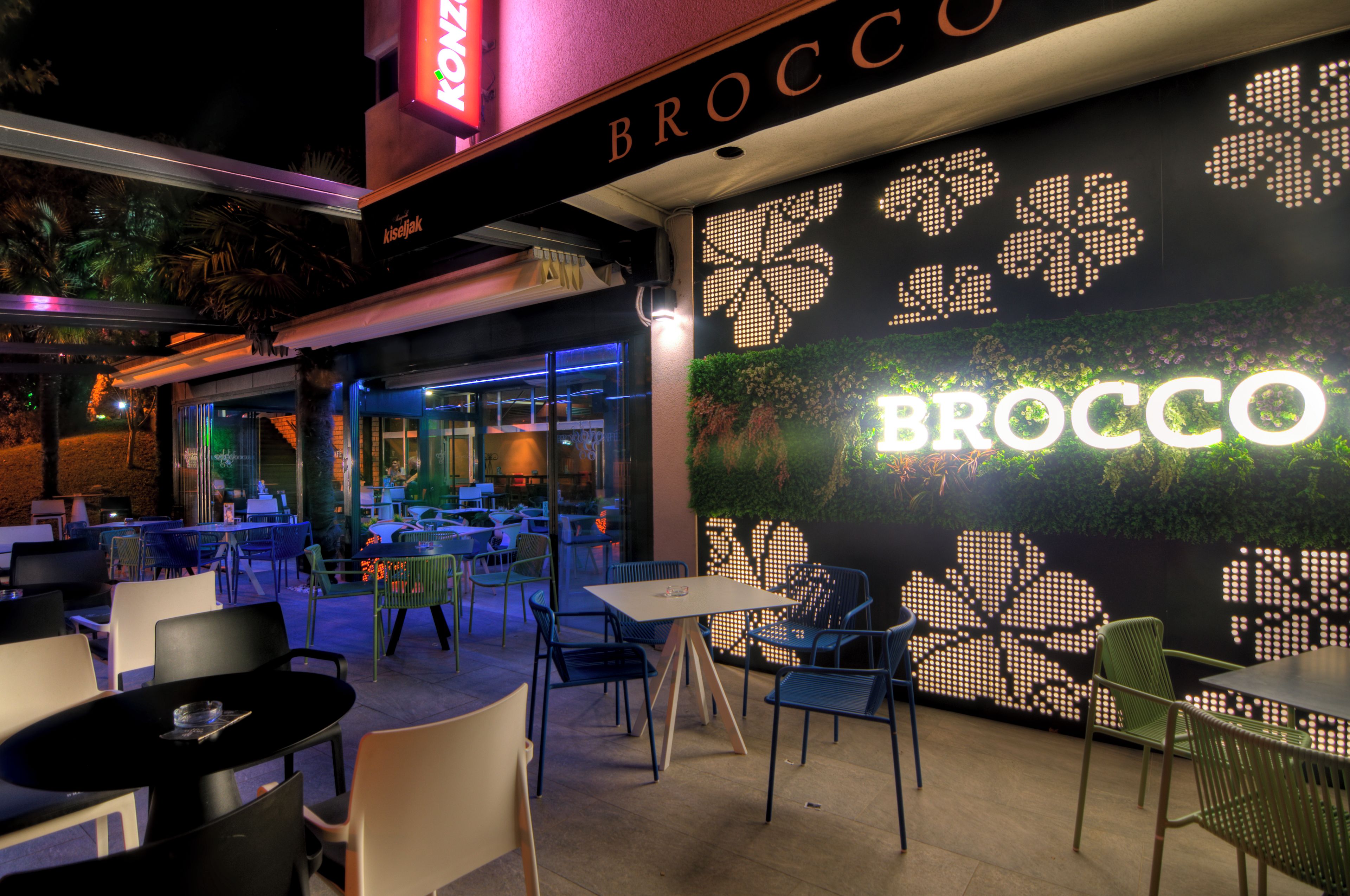 An outdoor dining area with tables and chairs, a wall with illuminated letters spelling 'BROCCO', and a neon sign above.