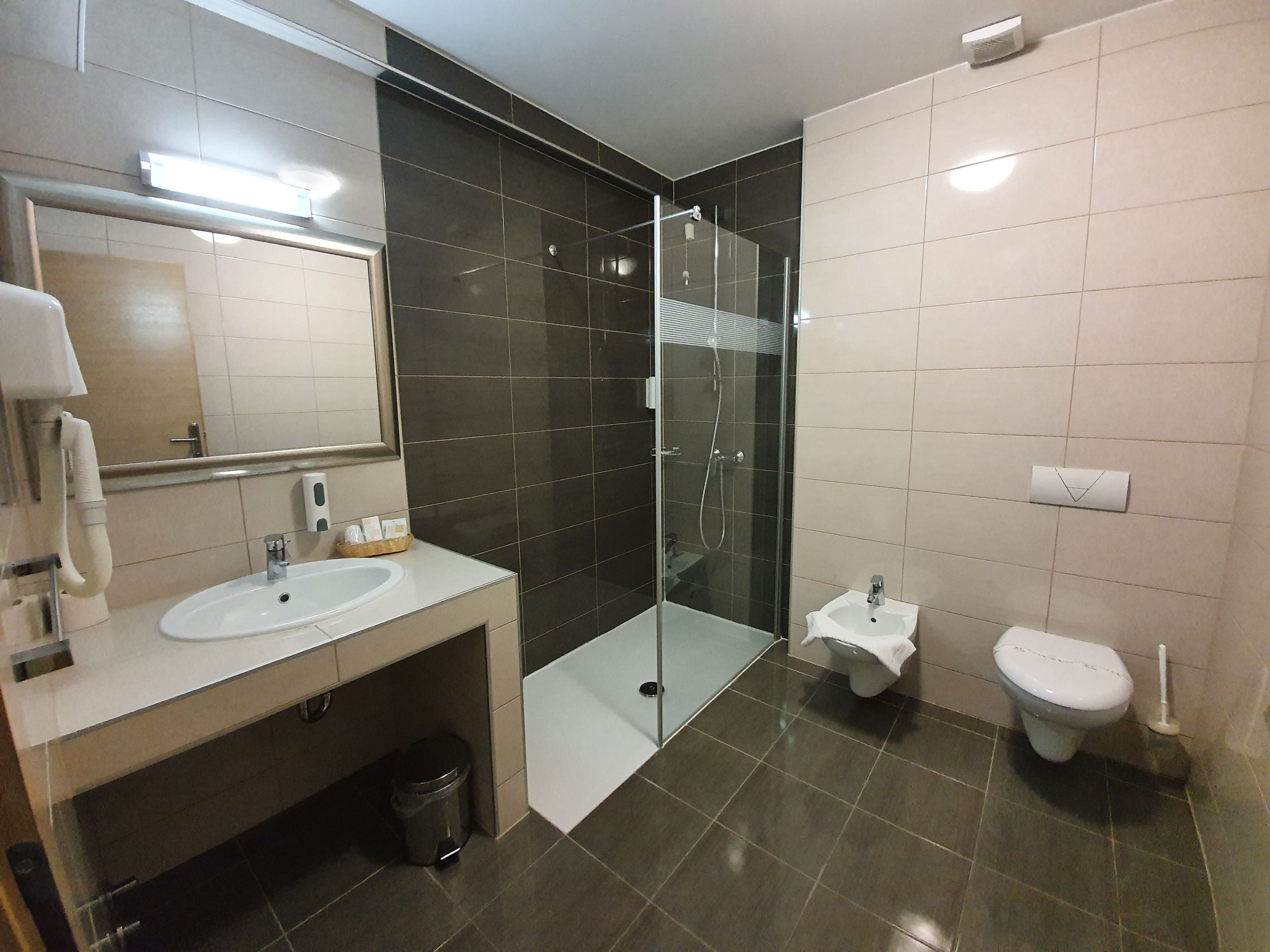 A modern bathroom with a glass-enclosed shower, two sinks, a toilet, and a trash bin.