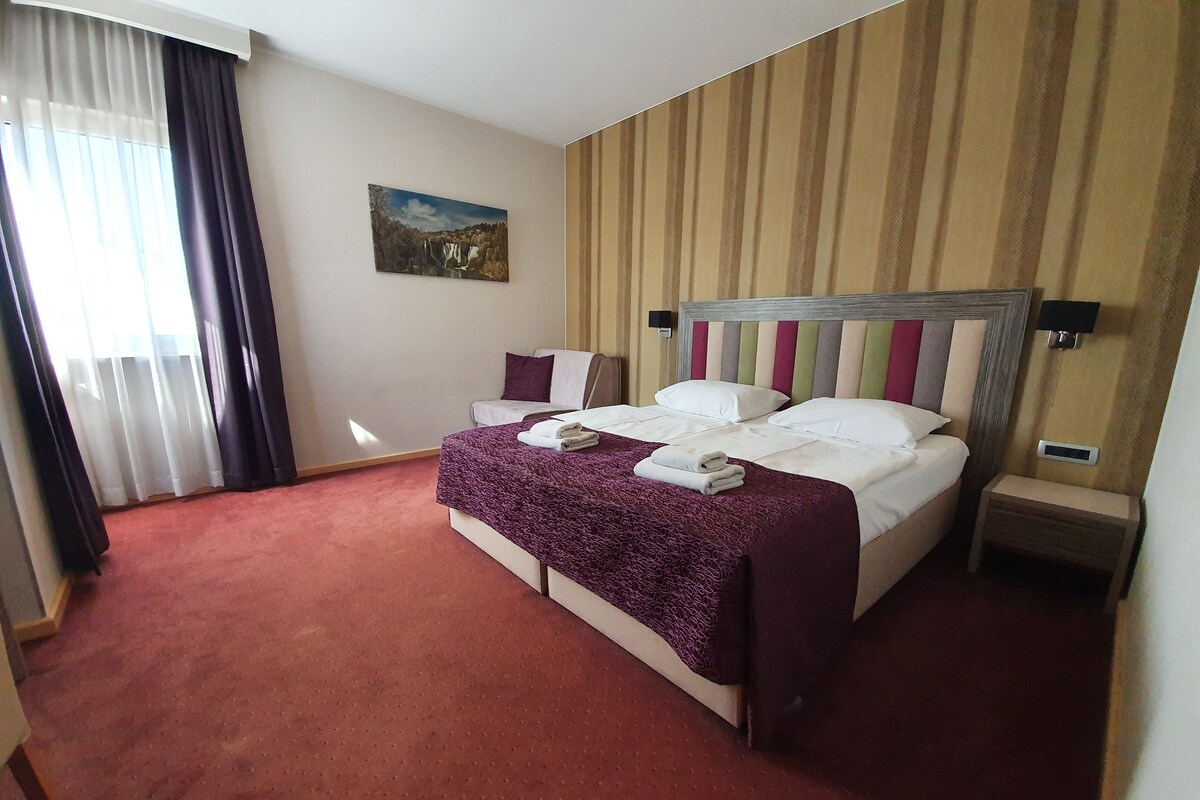 A hotel room with a large bed, a nightstand, a wall-mounted lamp, and a painting on the wall.