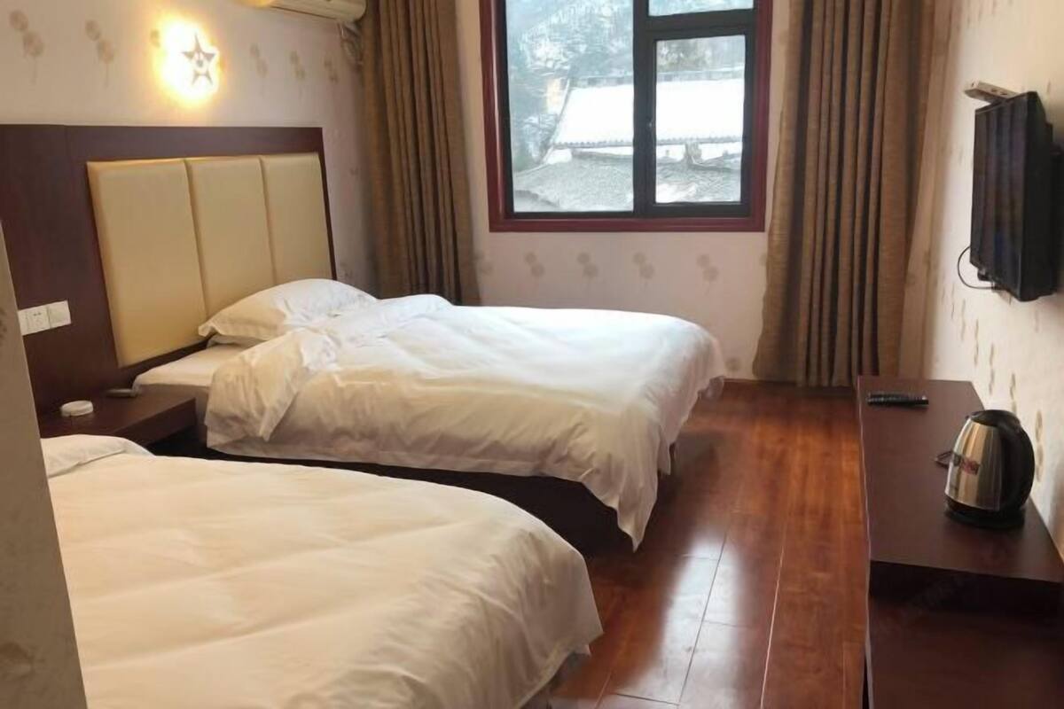 A hotel room with two beds, a wooden floor, a nightstand, a kettle, and a window with curtains.