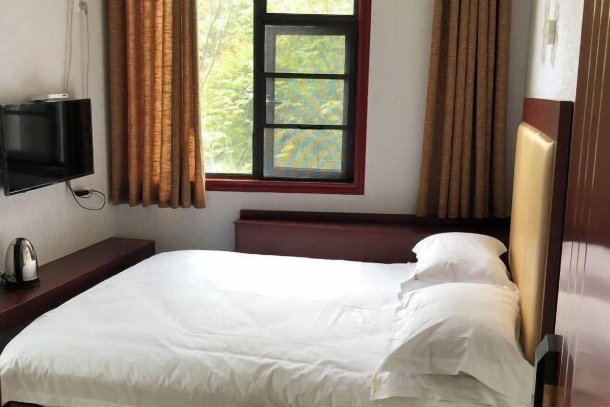 A neatly made bed with white linens, a bedside table with a kettle, a television mounted on the wall, and a window with curtains.