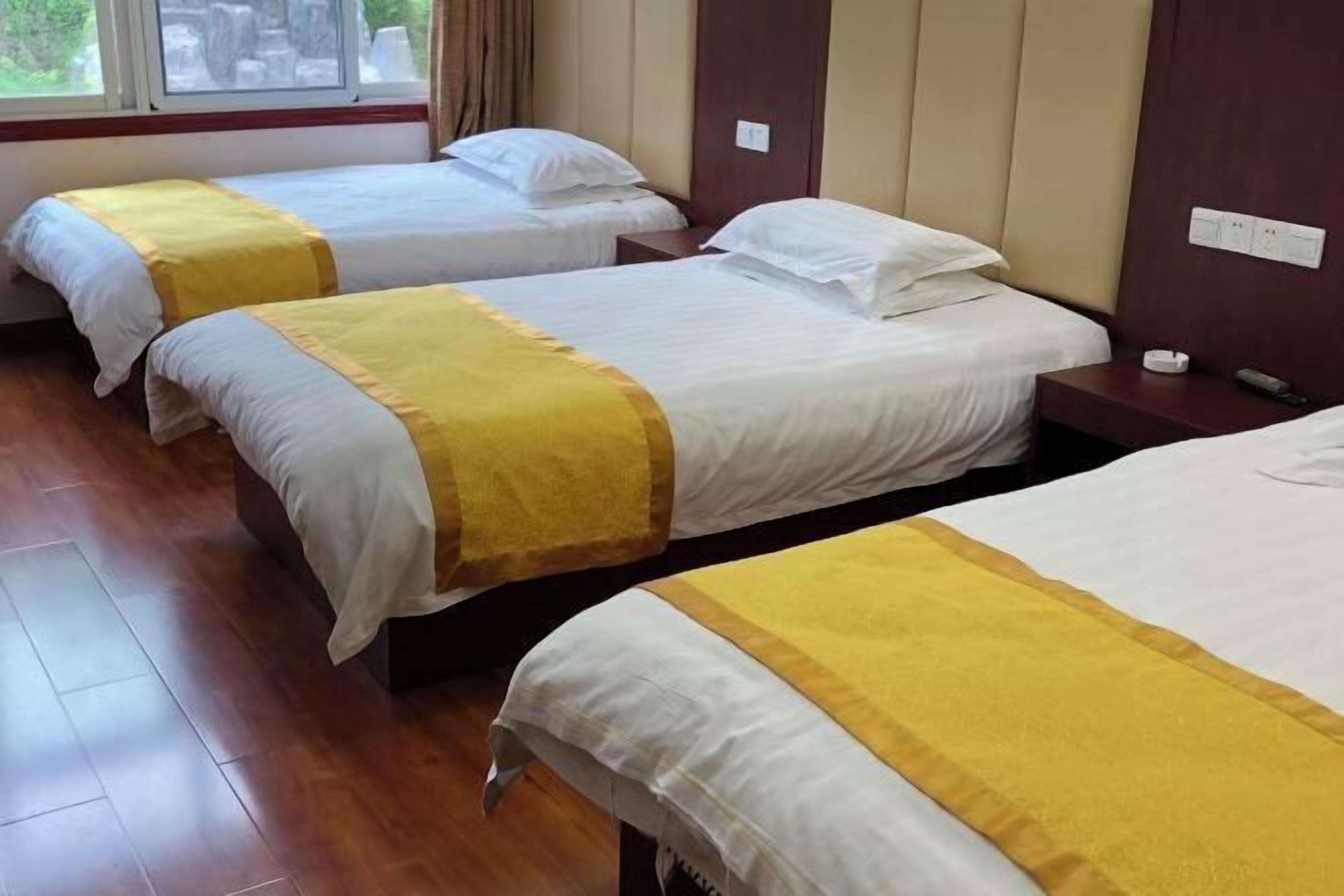A hotel room with three single beds, each with a yellow and white bedspread. The room has wooden flooring and a window with curtains.