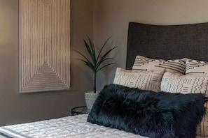 Un lit avec un oreiller noir et moelleux, une plante en pot et des coussins à motifs sur la tête de lit.