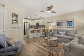 A modern living room with a grey sofa, a coffee table, and a clear glass table. There is a kitchen area with white cabinets and a bar stool. The room has a ceiling fan and wall art.