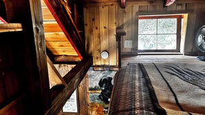 A wooden cabin interior with a bed, a window with a view of trees, and a wall-mounted fan.