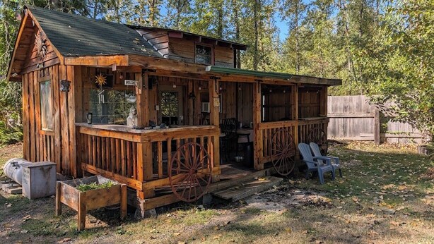 A rustic wooden cabin with a porch, a welcome sign, and a small outdoor seating area.