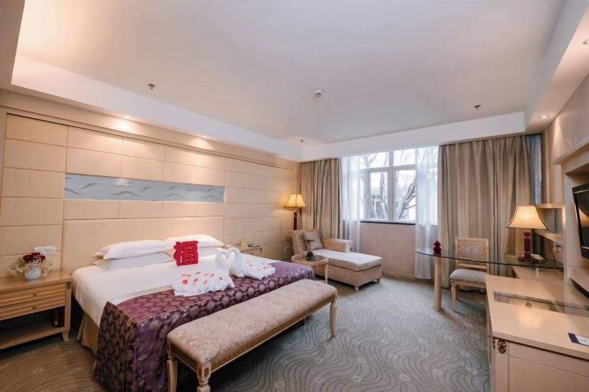 Ein Hotelzimmer mit einem Bett, einer Bank, einem Schreibtisch, einem Sessel, einem Fernseher und einem Fenster mit Vorhängen.