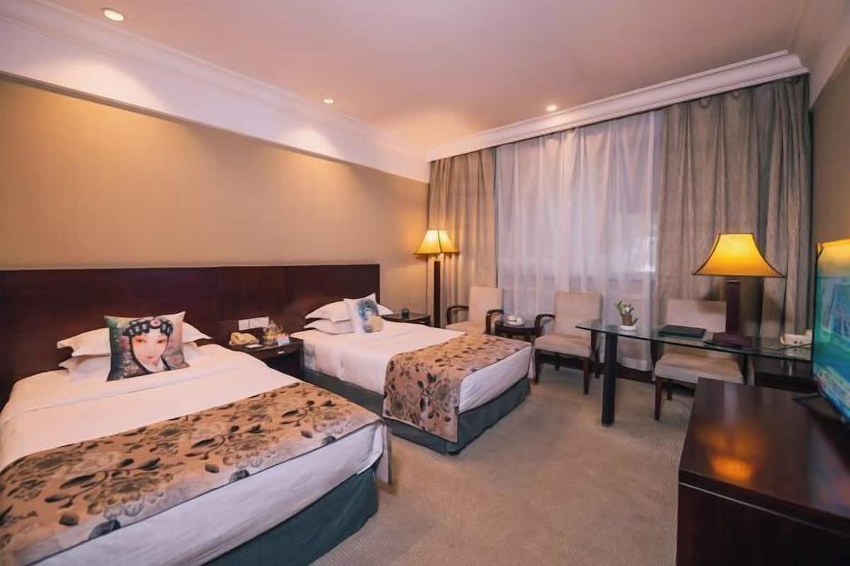 Ein Hotelzimmer mit zwei Betten, einem Schreibtisch, einem Stuhl, einem Fernseher und einem großen Fenster mit Vorhängen.