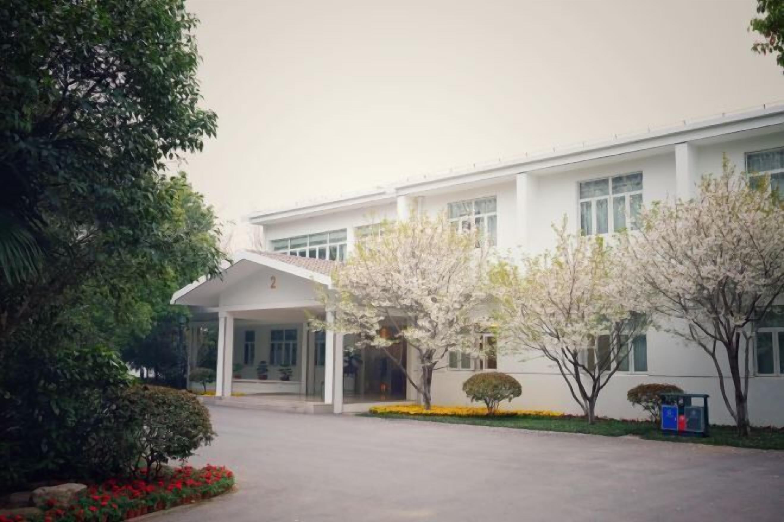 A modern building with a covered entrance, surrounded by greenery and flowering trees.